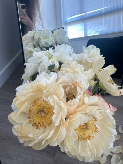 A close-up, eye-level, medium shot captures a bouquet of pale yellow and white peonies in a vase, positioned on a grey wooden floor. The flowers, with their soft, layered petals and prominent yellow stamens, dominate the foreground. To the left, a mirror reflects a woman holding a smartphone, her hand adorned with a ring. In the background, a dark object, possibly a chair, is visible, and beyond that, blinds cover a window, allowing light to filter through. The scene appears to be indoors, in a residential setting.