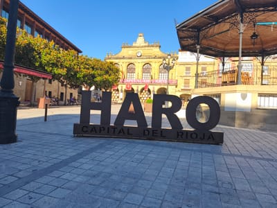 The image depicts a sunlit town square in Haro, Spain, with a prominent monument in the foreground. The monument, made of large, dark metal letters, spells out "HARO CAPITAL DEL RIOJA". Behind the monument, a historic building with arched windows and a clock tower is visible, adorned with banners. To the right, a decorative gazebo with a wooden roof and intricate metalwork stands. The square itself is paved with light-colored cobblestones. Several people are present, some seated on benches under trees, while others walk in the background. The overall scene suggests a pleasant day in a town square.