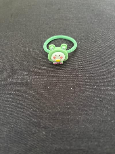 A cute, green frog-shaped hair tie is centered on a dark, textured surface. The frog has large, round eyes, a small yellow beak, and pink cheeks. Its body is green and appears to be made of soft material, possibly rubber or silicone. The hair tie itself is a thin, bright green elastic band. The background is a dark, likely fabric, surface with a subtle woven pattern. There are no people or any other objects in the image. The scene is a close-up shot, with the hair tie being the sole focus. There is no identifiable information that suggests a location in the city of Imeni Morozova, Russia.