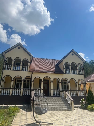 The image depicts a two-story cream-colored house with a brown tiled roof, situated in Bolshoy Molokish, Moldova. The house features a covered porch with arched openings supported by columns on the ground floor, and a balcony with similar arched openings and white balusters on the second floor. A staircase with tiled steps leads to the entrance. In the foreground, a hanging wicker chair with a gray cushion is visible. The ground is paved with interlocking tan and gray tiles. To the right of the house, a small evergreen tree and flowering bushes are present. The sky above is blue with scattered white clouds. Trees are visible in the background to the right. The scene appears to be an exterior view of a residential property on a sunny day.
