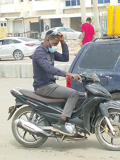 A street scene in Daurawa, Nigeria, displays a male individual seated on a black underbone motorcycle, identified by the visible "Typhoon" branding. The subject wears a dark blue long-sleeved collared shirt, grey trousers, black socks, and black sneakers. A light blue disposable face mask covers his nose and mouth. He has sunglasses pushed onto his head and is observed adjusting them with his right hand; his left hand grasps the left handlebar grip. To the immediate right, a dark blue vehicle, consistent with an SUV or van, is partially visible, featuring a yellow container secured to its roof. In the background, another male figure, dressed in a red garment, walks away from the camera. The ground consists of a dusty, light-colored surface, and concrete barrier blocks are positioned along a median. Further back, multiple light-colored buildings, some appearing unfinished, line the street, interspersed with parked silver sedans.