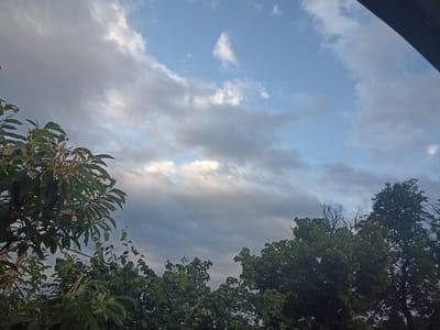 The image shows a sky with clouds and trees in the foreground. The sky is a mixture of blue and gray, with some white clouds scattered throughout. The trees are lush and green, with some of them obscuring parts of the sky. The time of day appears to be late afternoon or early evening, as the light is soft and diffused. The weather seems to be fair, with a few clouds but no indication of rain. The scene is likely a natural outdoor setting, possibly a park or a backyard, given the presence of trees. There is no visible text in the image.