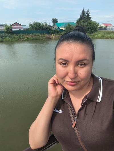 A woman with dark hair pulled into a bun is captured in a medium-close-up selfie against a backdrop of a river and residential buildings. The woman is wearing a brown polo shirt with white trim and a zipper. Her left hand is resting on her chin, and she is looking towards the camera. In the background, a body of water is visible, with a grassy bank and several houses with colorful roofs and fences. Two tall pine trees stand out among the greenery. The sky is partly cloudy. The overall impression is a casual, outdoor portrait. The location is identified as Sokury, Russia.