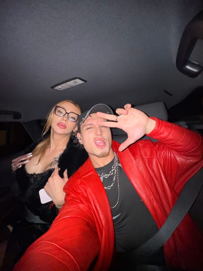 The image depicts two individuals, a female and a male, inside the passenger compartment of a vehicle, specified as located in Asnières-sur-Seine, France. The scene is illuminated by an internal light source, suggesting a low-light or nighttime setting.
The female subject is positioned on the left, slightly behind the male. She has light-colored hair, pulled back into a ponytail, and wears dark-rimmed eyeglasses. Her mouth is slightly open, with her tongue partially visible. She is dressed in a black garment, possibly lace or sheer, with a plunging neckline, covered by a black faux fur or feathered wrap. Her hands display rings and long, white manicured nails; her right hand holds a light-colored mobile phone against her chest.
The male subject is in the foreground, on the right. He wears a black baseball cap backward, a red leather jacket over a black t-shirt, and multiple silver-toned chains around his neck. He has a goatee, an open-mouthed expression with his tongue visible, and squinting eyes. His left hand is raised, forming a peace sign (V-shape). Multiple earrings, including hoops and a cross-shaped drop, are visible on his ears, and he wears rings on his left hand. A black seatbelt crosses his right shoulder and chest.
The vehicle's gray headliner is visible at the top of the frame, along with a rectangular overhead light fixture. Both individuals are looking towards the camera, suggesting a self-portrait (selfie).