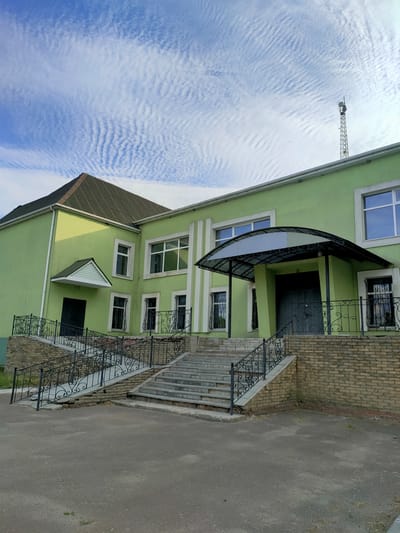 A wide, low-angle shot captures the front exterior of a light green, two-story building under a bright blue sky with scattered, wavy white clouds. The building has a dark green, sloped roof with white gutters. A prominent feature is a set of wide, gray concrete stairs leading up to a covered entrance. The entrance is protected by a dark, arched awning with a metal frame and translucent panels. Double dark doors are visible beneath the awning.

Flanking the stairs are decorative black metal railings with intricate scrollwork. The base of the stairs and the walls of the building's foundation are constructed of light brown brick. On the left side of the building, a smaller, dark green door is situated at the bottom of a short set of stairs with similar metal railings. The building's facade features several rectangular windows with white frames, some with black metal bars. The upper windows are larger and appear to be office or classroom windows. White decorative vertical trim elements are visible on the right side of the building.

To the right of the building, a tall, slender metal tower with antennae is visible against the sky. The scene appears to be a public building or institution. The lighting suggests it is daytime, likely morning or afternoon, with the sun casting shadows on the left side of the building. The overall impression is one of a well-maintained, functional building in an outdoor setting. No people are visible in the image. The weather appears to be clear and pleasant. The setting is urban or suburban, with a paved area in front of the building.