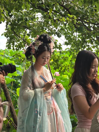 In Xihu District, China, two young women are photographed in a natural setting with lush green foliage. The woman on the left is dressed in traditional Chinese attire, a flowing light blue and pink gown, adorned with flowers in her hair. She holds a small blue fan in her hands. To her right, another woman, wearing a pink t-shirt, looks down and smiles. In the foreground, a camera lens is visible, suggesting a professional photoshoot is underway. The background is dominated by large green lotus leaves and a single pink lotus flower, indicating the scene is likely near a pond or lake. The sunlight filters through the leaves, creating a soft, diffused lighting.