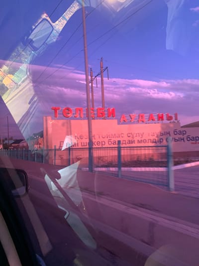 The image is a double exposure photograph taken from inside a vehicle, looking out at a sunset scene. The primary focus is a large red sign with Cyrillic lettering, which reads "ТОЛЕБИ АУДАНЫ". Below this, there is another line of text, also in Cyrillic, which appears to be a slogan. The sign is pinkish in color and is situated in front of a pale pink building. A metal fence runs along the front of the property.
The overlaid image shows the interior of a car, including a rearview mirror and a reflection of the sky on the windshield. The sky is a gradient of purple and pink, with some clouds visible. Power lines and utility poles are also present in the scene. The overall impression is a somewhat surreal and artistic representation of a place in Kazakhstan.
The red lettering on the sign is "ТОЛЕБИ АУДАНЫ", which translates to "Tolebi District". This indicates the location is within the Tolebi District of Kazakhstan. Given the context of Tolebi District, and that Tolebi is a district in the Turkistan Region of Kazakhstan, the specific location is likely the administrative center or an entrance to the Tolebi District. The image captures a roadside scene at dusk, with the sign serving as a landmark.