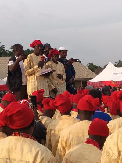 This image captures a large outdoor gathering in Omicha Umu Awada, Nigeria, likely a cultural or community event. The central focus is a group of men standing prominently, possibly on a stage or raised platform, addressing an audience.
In the midground, a central figure, dressed in a traditional light-colored, patterned garment and a red knitted cap (a significant cultural symbol in Igbo land), is speaking into a microphone while holding a document or book. To his right, an older man, similarly dressed in a light garment and red cap, also holds a document. Another man in a white t-shirt and dark vest stands to the left, holding a microphone, seemingly assisting or co-hosting. Behind these main speakers, several other men are visible, some holding smartphones or cameras to record the event. One man wears a white baseball cap.
The foreground is filled with the backs of numerous attendees, primarily men, all dressed in similar traditional light-colored, long-sleeved garments and distinctive red knitted caps, some with a pom-pom. Many in the audience are holding up smartphones, capturing the moment. One man in the immediate foreground wears a red beaded necklace.
The setting appears to be an open field or community ground, with buildings featuring corrugated iron roofs and trees visible in the background. A large white tent is also present in the background, partially obscuring text that appears to read "ADOUNA REA," possibly part of a company name like "ADONAI REAL ESTATE." The lighting suggests late afternoon or early evening, with a bright but soft sky. The event appears to be well-attended and significant, given the traditional attire and the number of people recording.