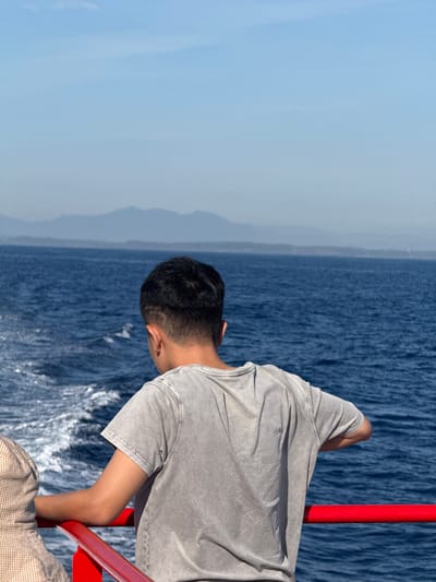 The image shows a view from the deck of a boat on the ocean. In the foreground, two people are visible. The person on the right is a young male, viewed from behind, wearing a light gray t-shirt. His right arm is resting on a bright red railing. To his left, partially visible and also leaning on the railing, is another person wearing a patterned garment, possibly a hat or clothing. The water is a deep blue, with white wakes trailing behind the boat, indicating movement. In the background, a hazy blue horizon separates the sea from the sky. Distant, muted mountains are visible on the horizon. The sky is a pale blue with a few faint clouds. This scene could be depicting a ferry ride or cruise in the waters near Dong Son, Vietnam, a coastal area known for its beaches and sea access.