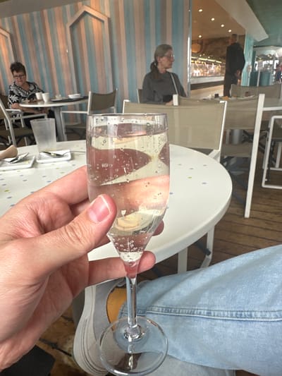 An indoor scene in a dining establishment in Puerto del Rosario, Spain, is depicted. In the immediate foreground, a light-skinned hand, appearing male, grips the stem of a clear champagne flute filled with a sparkling, transparent liquid exhibiting numerous effervescent bubbles. The individual holding the glass wears light blue denim jeans and a grey sneaker with a yellow-tan interior is partially visible. The glass is positioned over a white circular table adorned with small blue, green, and yellow polka dots. White rectangular dishes and silver cutlery are visible on this table, along with a clear plastic drinking glass. In the mid-ground to the left, a middle-aged woman with short, dark hair and glasses is seated at a separate table, wearing a black and white patterned top. A white mug and other dishes are on her table. The wall behind her features vertical stripes in pastel blue, pink, and white, with a white decorative arch-shaped frame. To the right, another middle-aged woman with long, light brown hair tied back and wearing glasses and a grey long-sleeved shirt is seated at a table. A dark object hangs from her shoulder or rests on her table. Further in the background, to the right, a man in a dark shirt stands with his back partially turned, positioned near an open area with a visible refrigerated display case containing various food items, indicative of a deli or buffet section. Recessed lighting is visible on the ceiling in this area. The flooring throughout the establishment consists of light brown wooden planks. White and beige-colored chairs are distributed across the scene.