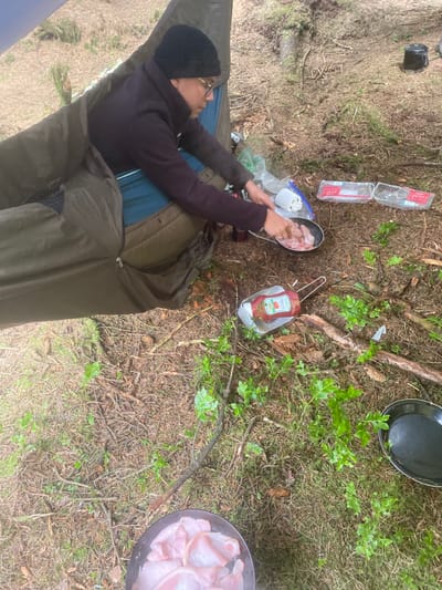 A person wearing a black hat and a brown sweater is sitting inside a green hammock in the woods. They are cooking something over a small camping stove. There are two black frying pans nearby, one containing raw meat and another containing some oil. There is a bottle of ketchup on the ground. The scene is set in the forest, which is covered in pine needles and branches. There is a small amount of green undergrowth around the base of the trees. The person is seated in the middle of the frame, facing right. The hammock is positioned off to the left of the image. The person is using their right hand to stir the contents of the frying pan. The setting is likely in Macclesfield Forest, United Kingdom, based on the provided latitude and longitude.