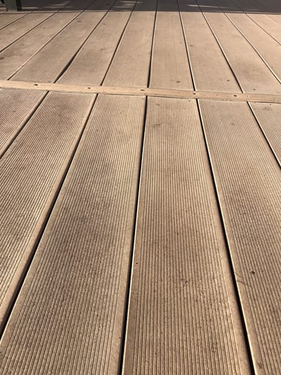 The image shows a close-up, angled view of a light brown wooden deck. The deck is made of parallel planks, each with a textured surface featuring fine vertical grooves. A horizontal wooden strip, secured by visible screws, crosses the deck about one-third of the way from the bottom of the frame. The deck appears to be outdoors, with dappled sunlight and shadows suggesting it is near a structure or trees. There is some dirt and debris scattered on the planks. The scene is devoid of people. The location is identified as Azov, Russia.