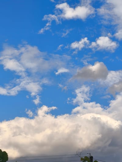 This is a low-angle, vertical, medium shot of a partly cloudy sky above Beșghioz, Moldova. The sky is a vibrant blue, dotted with various white and gray clouds. The clouds vary in size and density, with some appearing wispy and thin, while others are larger and more substantial. In the foreground, silhouetted against the sky, are parts of a utility pole with wires and a leafy green vine. The lighting suggests it is daytime, with the sun illuminating the clouds from above. The overall impression is one of a typical summer sky in the region.