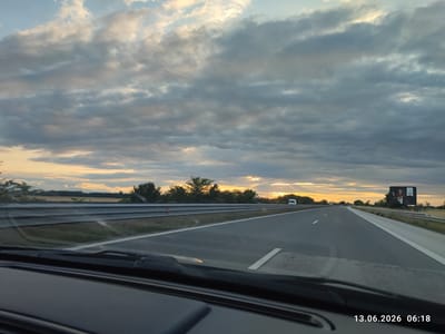 The image captures a view from inside a vehicle traveling on a highway, likely on the outskirts of Tsaratsovo, Bulgaria. The perspective is from the driver's seat, looking forward through the windshield, which shows some dust and wiper streaks. The highway stretches ahead, with multiple lanes marked by white lines. Guardrails are visible on the left side of the road.
The sky is filled with dramatic, cloudy formations, with patches of sunlight breaking through, suggesting either sunrise or sunset. The warm hues of the setting or rising sun are visible on the horizon, casting a golden glow. A billboard is present on the right side of the highway, featuring an advertisement with an image of a person. In the distance, several vehicles, including a white van, are driving on the highway. The landscape flanking the road appears to be rural, with fields and trees lining the horizon. The timestamp "13.06.2026 06:18" is overlaid on the bottom right corner of the image.