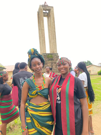 In Wang, Nigeria, a group of young people are gathered outdoors, possibly for a cultural event or celebration. Two young women are prominently featured in the foreground, both smiling and looking towards the camera. They are dressed in vibrant, traditional attire.
The woman in the front left wears a green and yellow striped wrap over a black t-shirt, adorned with a colorful beaded necklace and matching headband. Her hair is styled in a traditional wrap. The woman behind her, slightly to the right, wears a red and green striped sash over a black t-shirt, with a similar beaded headband and necklace. Her hair is in long, dark braids. Another young woman in the background, also in traditional attire, is partially visible.
The setting appears to be a grassy area with a concrete structure, possibly a pillar or monument, on the left side of the frame. A building with an orange roof and windows can be seen in the background, suggesting an outdoor gathering. The sky is overcast, indicating a cloudy day. The overall mood is festive and joyful, with the subjects appearing relaxed and happy. Some of the visible text on the t-shirt of the woman in the front right reads: "EAT SLEEP CLINICAL @ PANIC".