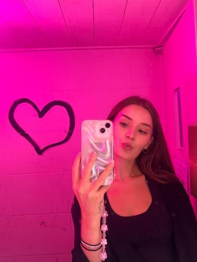This image captures a mirror selfie of a female subject within an intensely magenta-illuminated indoor environment. The subject, appearing to be in her mid-20s to early 30s with long brown hair, is depicted from the chest up. She wears a dark top, a dark jacket, small hoop earrings, a delicate necklace, and a nose stud. Her right hand holds a smartphone with a pearlescent, wavy-patterned case and a decorative beaded phone charm featuring white and pink flower-shaped beads. Her lips are pursed in a 'kiss' gesture, and her gaze is directed towards the phone's camera as she captures her reflection. The background consists of a wall and a paneled ceiling, both saturated with vibrant pink light. A large, black, hand-drawn heart symbol is visible on the wall to the left of the subject. To the right, a vertical illuminated panel emits pink light, and a small, dark object, possibly a security camera or sensor, is mounted high in the corner of the room. The exact location within Mapo-gu, South Korea, cannot be determined from the visual content of the image.