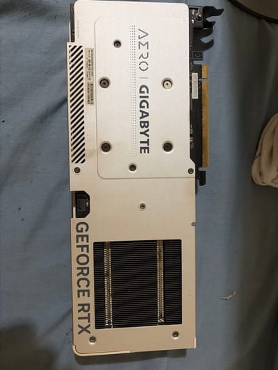 A Gigabyte AERO GeForce RTX graphics card sits on a blue fabric surface. The card features a silver backplate with the AERO and Gigabyte logos, along with visible screws. A central rectangular section exposes the heatsink fins. "GEFORCE RTX" is printed along the bottom edge. A sticker on the backplate displays the model number, "GV-N4060AERO OC-8GD," and the serial number, "SN240541006455". The text "BIOS OC/SILENT" is printed on the side.
