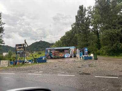 The image depicts a roadside food truck stand on a cloudy day, likely located in or near ნიგოითის თემი, Georgia, given the context. The food truck is colorful and adorned with various graphics, suggesting it sells snacks or drinks. In front of the truck, there are blue benches, some white chairs, and a green trash can. A signpost with text in a foreign language, possibly Georgian, is visible to the left of the food truck, along with what appears to be a smaller building behind it. The ground is wet and appears to be asphalt or gravel, with puddles of water, indicating recent rain. Trees and lush green hills form the background, suggesting a rural or semi-rural setting. There are no people visible in the image.