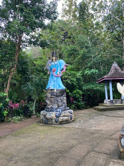 A concrete statue of a woman in a blue dress and a crown stands on a round, multi-level base decorated with a multi-colored dragon in the foreground. The woman holds a pink rose in her right hand. Behind the statue is a small, open-air pavilion with a white, tiled floor. There are trees and bushes surrounding the structures and a dirt path leads to the statue. The location is in the city of Kecamatan Manyaran, Indonesia, at the coordinates -7.826, 110.816.