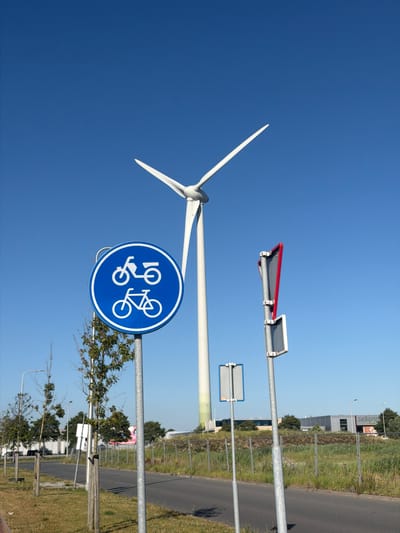 This image captures an outdoor scene under a clear blue sky, suggesting a bright and sunny day. The primary subject is a wind turbine, with its white blades visible against the sky, indicating a relatively calm wind condition as the blades are not in rapid motion.
In the foreground, a blue circular traffic sign with white pictograms is prominently displayed on a metal pole. The sign depicts two bicycles, one with a rider and the other empty, suggesting a shared bicycle path or a warning related to cycling. This is a common sight in areas with designated cycling routes.
Further back, a paved road runs parallel to a grassy area with some trees and industrial or commercial buildings in the distance. The presence of the wind turbine and the road suggests an area that might be a mix of rural and industrial, possibly on the outskirts of a town or in a designated energy production zone. The buildings have a modern, functional appearance.
The overall impression is one of a functional, modern infrastructure scene, likely in the Netherlands given the location context provided, where renewable energy and cycling are often integrated into the landscape. The lighting indicates it's daytime, likely morning or afternoon, with no indication of specific weather beyond clear skies. There is no visible text on the wind turbine or the natural elements. The traffic sign is purely symbolic.