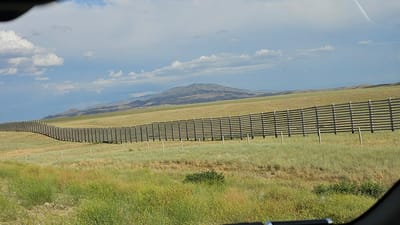 An exterior, eye-level, wide shot captures a wooden fence that stretches across a grassy plain towards rolling hills and distant mountains under a partly cloudy sky. The fence, constructed of horizontal wooden planks supported by vertical posts, curves gently from the left foreground into the distance. The plain is covered in a mixture of green and dry grasses, with some scattered yellow wildflowers in the foreground. In the background, the landscape transitions to softer, muted tones of hills and mountains, culminating in a prominent, larger mountain in the center-left of the frame. The sky features scattered white and grey clouds against a blue backdrop, with a faint contrail visible in the upper right quadrant. The overall scene depicts a rural, open landscape.