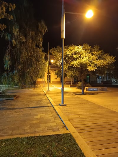 This is a night-time photograph of an outdoor paved area, likely a plaza or promenade, illuminated by streetlights and architectural lighting. The ground is paved with light-colored rectangular stones on the left and a wooden boardwalk with parallel planks on the right. A well-maintained patch of green grass is visible in the foreground. Several streetlights on tall poles are present, casting a warm yellow light. One streetlight in the upper right corner is particularly bright.

To the left, a weeping willow-like tree with long, drooping branches dominates the scene, its foliage illuminated by the artificial light. Further back, a building with arched doorways and windows is visible, suggesting it might be a public building or commercial establishment. Trees with fuller canopies are scattered throughout the area, also lit by the streetlights. Some benches, one of which is made of concrete with a slatted metal insert, are present in the background. The overall atmosphere is serene and quiet, typical of an urban area at night. This scene is located in the city of Roseto degli Abruzzi, Italy.