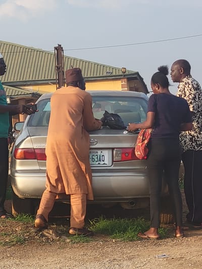 In Kwamba, Nigeria, a group of people stand near the trunk of a silver Toyota car. A man in a brown traditional robe and cap is positioned at the trunk, appearing to interact with it. Two other people, a man in a patterned shirt and a woman in a blue shirt and a red bag, are also near the trunk. The car's license plate, "NYIA 63AQ", is visible. There is green grass along the roadside, and a building with a green roof is in the background.