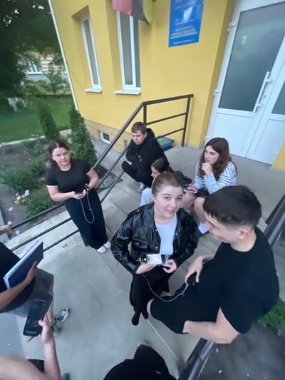 A group of young people are gathered on concrete stairs in front of a yellow building, likely a school or public institution. The building has white-framed windows and a white door. The stairs have a black metal railing. Some individuals are sitting on the stairs, while others are standing or leaning. One person in the foreground is holding a phone and a blue folder. Another person, wearing a black leather jacket, is holding a phone. The environment appears to be outdoors with some greenery and trees visible in the background. The overall atmosphere suggests a casual social gathering.