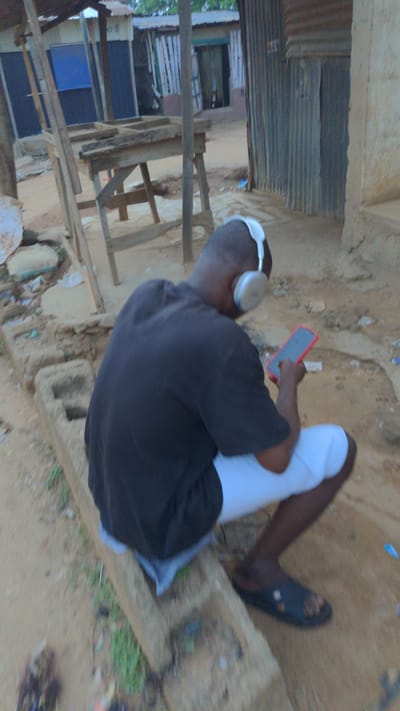 A person is seated outside near a building constructed of corrugated metal and lumber. The individual is wearing white pants, a black shirt, and a black sandal on one foot. They have headphones on and are holding a red phone. The surrounding environment appears to be a sandy area. The location is Masaka, Nigeria.