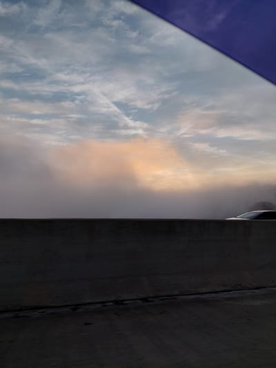 The image is a vertical, wide-angle shot of a cloudy sky. A portion of a dark, textured surface, possibly a wall or structure, runs along the right side of the frame. A sliver of a white vehicle with a dark window is visible at the top right edge. The sky is filled with a mix of white and grey clouds, with streaks of orange and yellow light suggesting sunrise or sunset. The lower left corner of the image shows a sliver of a dark purple surface, which could be an awning or fabric. The overall impression is of an outdoor scene, possibly urban, with a dramatic sky. The time of day is likely dawn or dusk due to the colors in the sky. No text is visible in the image. The location is provided as Richmond, United States.