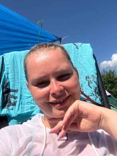 The image is a close-up, selfie-style shot of a young woman with blonde hair pulled back, facing the camera and smiling. She is outdoors on a sunny day, with a clear blue sky in the background. A blue tarp is visible behind her, along with a light blue towel featuring an elephant print. A small green sprout is growing from the top of her head, seemingly attached to something unseen. She is wearing a light-colored hooded shirt. Her right hand is raised, with her fingers gently touching her chin. The scene appears to be at a campsite or outdoor gathering, as indicated by the tents and natural surroundings in the background. The location is specified as the city of Жемчужный, Russia.
