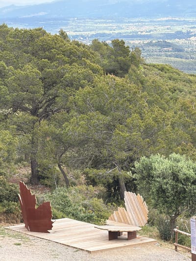 The image depicts a scenic overlook with a wooden observation platform located in Torrelles de Foix, Spain. The platform features a metal sculpture shaped like a leaf. The scene overlooks a vast landscape with rolling hills and vegetation. The sky is partially visible in the distance.