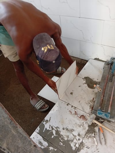 An unidentified dark-skinned man wearing shorts, sandals, and a cap is installing white marble-patterned tiles on a floor. The work is taking place indoors, near a wall also tiled with white marble-patterned tiles. A trowel, tile cutter, hammer, and pliers are present on the tile-covered floor. The location is in the city of Masaka, Nigeria.