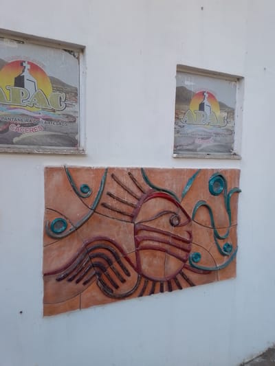 The image shows a white wall with two framed posters and a relief sculpture. The posters are located in rectangular recesses in the wall and appear to be advertisements or informational signs. The relief sculpture is mounted on the wall below the posters and depicts an abstract design. The sculpture is made of terracotta-colored material and features raised elements in red and blue. The overall scene suggests an outdoor location, possibly a public space in the city of Cáceres, Brazil, given the text visible on the posters, which includes "CACERES". The lighting suggests it is daytime.