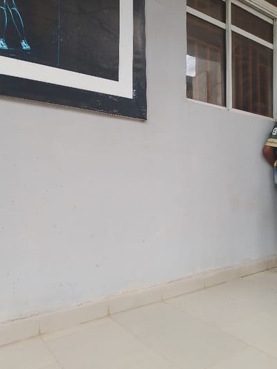 The scene appears to be an exterior shot, possibly along a building in Ogboko, Nigeria. The wall is painted a light gray color and has a large, framed picture on it. A window is visible to the right of the picture. In the lower part of the frame, there are light-colored tiles on the floor. A person is partially visible on the right side of the frame. The image focuses on the building's features, providing a simple documentation of the external facade.