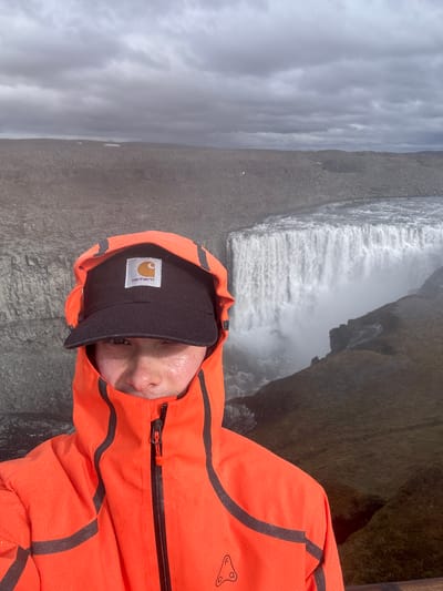A vertical, slightly overhead shot captures a person near a powerful waterfall under an overcast sky. The subject, in the lower right foreground, wears a bright orange waterproof jacket adorned with dark grey or black stripes and a black baseball cap. Numerous water droplets are visible on the jacket, indicating mist from the waterfall or rain. The person's face is partially seen beneath the brim of the cap, looking upwards towards the camera.

In the background, a wide, intense waterfall plunges over a dark, rocky cliff, generating significant mist at its base. The surrounding landscape features rugged, dark rock formations and sparse vegetation. The sky is entirely covered by thick, grey clouds, creating a subdued natural light.

The scene suggests an outdoor adventure in a wild, majestic environment, consistent with the provided location context of Iceland. The time of day is daylight, characterized by the cloudy conditions.

Visible text includes the "Carhartt" logo on a white patch on the front of the black baseball cap. Additionally, a distinct black logo, possibly a stylized figure or emblem, is present on the upper right arm/shoulder of the orange jacket.