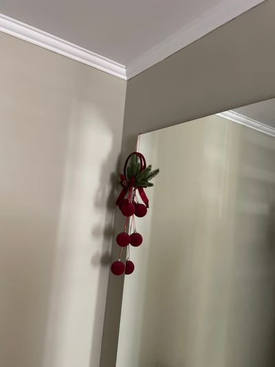 The image displays a Christmas decoration hanging from a corner where two walls meet. The decoration consists of greenery, red ribbons, and several red baubles. The walls are painted a neutral, light color and meet at a white crown molding. The lighting in the scene creates shadows on the walls, suggesting an indoor environment. There are no people visible in the image. The specific location in Cahul, Moldova, cannot be determined from the visual information provided.
