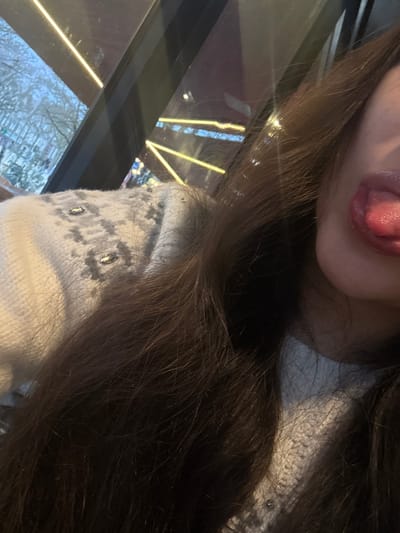 A person sticks their tongue out in a selfie. Long, dark brown hair dominates the image. The individual wears a white sweater or garment with a fair isle pattern in gray, accentuated by decorative studs or beads. Reflections of light and a cityscape can be seen through a window beside the person.