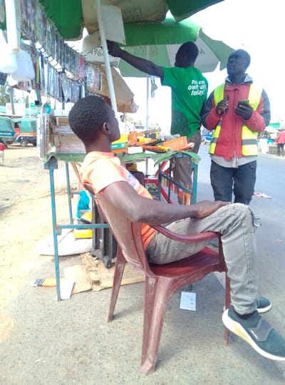 An outdoor, daytime scene in South Kabras ward, Kenya, depicts three adult males near a roadside stall. In the foreground, a male with short, dark, curly hair is seated on a dark red plastic chair, facing right and slightly upwards. He wears an orange t-shirt, light grey ripped jeans, and dark sneakers with white soles. His left arm rests on his left thigh. To the immediate right, a makeshift stall with a green umbrella features numerous plastic-packaged items, likely mobile phone accessories, hanging from a display rack. White plastic bottles also hang from the stall. Various items, including what appear to be tools and an orange broken plastic tray, are arranged on the stall's surface. A male individual, facing away from the camera, stands behind the stall, wearing a green t-shirt with partially visible white text "Pay as, own way today!" and reaches his right arm upwards towards the umbrella or hanging merchandise. To the right of the stall, a third male stands, facing left, wearing a red jacket with a bright yellow reflective vest over it, dark trousers, and dark gloves. He holds a dark smartphone in his left hand and his right hand is raised. The ground is a mix of light-colored dirt and asphalt, with pieces of cardboard under the stall and an orange-handled utility knife lying near the seated individual's feet. In the background, other vehicles, including a green tuk-tuk-like vehicle, are visible on a road under a bright sky.