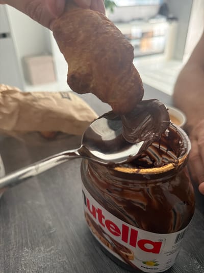 A close-up shot shows a person's hand holding a croissant, which is being dipped into a jar of Nutella. The jar is predominantly brown with a white label featuring the red "Nutella" logo. A silver spoon is also visible, scooping the chocolate-hazelnut spread from the jar. The background is blurred, but appears to be an indoor setting with a grey, textured surface, possibly a table. A crumpled brown paper bag is also visible in the background. The lighting suggests it could be daytime. No specific weather or emotional cues are evident. The visible text on the jar includes "Nutella" and "Ferrero".