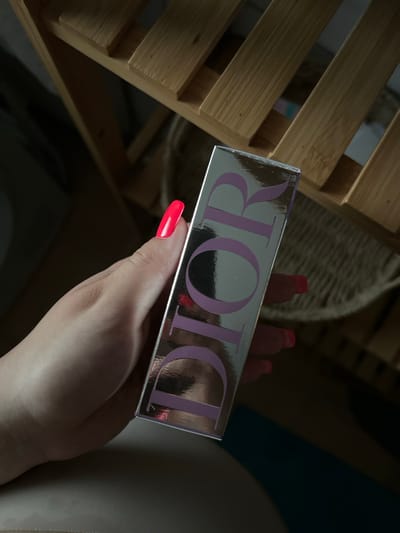 A hand with bright pink, long acrylic nails holds a silver rectangular box with the word "DIOR" printed vertically in lavender lettering. The box is angled towards the viewer and is partially in focus. In the background, a light-colored wooden slatted shelf is visible, with some dark areas suggesting it is placed in a room. The overall lighting suggests an indoor environment. No specific location in Waldbröl, Germany can be determined from this image.