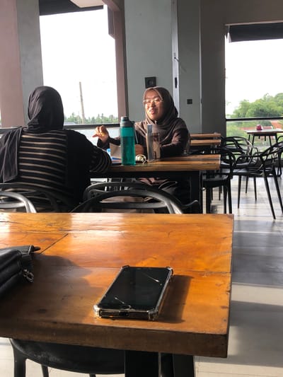 Two women are seated at tables in what appears to be an outdoor or semi-outdoor cafe. The woman on the left is wearing a black hijab and a striped top, with her back to the camera. The woman on the right is wearing a brown hijab and glasses, facing left and engaging in conversation with the other woman. She is holding a blue water bottle. A smartphone with a cracked screen and a black handbag are on the wooden table in the foreground. The background shows trees and buildings, suggesting an urban or suburban setting in Geulumpangsulu Timu, Indonesia.