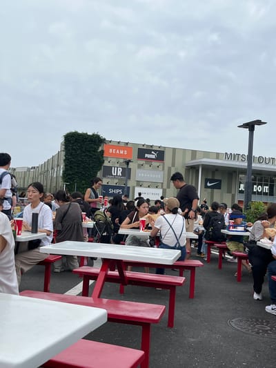 The image depicts an outdoor dining area at the Mitsui Outlet Park Iruma, located in Iruma, Japan. The scene is filled with numerous people, mostly Asian, seated at red and white picnic tables, eating and socializing under an overcast sky. The architecture of the outlet mall is visible in the background, featuring large signs for brands such as BEAMS, UR URBAN RESEARCH, SHIPS, PUMA, ABAHOUSE, AMETROPE, NIKE, and MITSUI OUTLET. People are dressed casually in t-shirts, shorts, and jeans. Some individuals are holding red cups, suggesting they are consuming beverages. Children are also present, some being held by adults and others seated at tables. The ground is asphalt, and a manhole cover is visible in the lower right portion of the image. A tall black lamppost stands to the right of the frame. The overall atmosphere appears to be that of a busy, casual public gathering place.