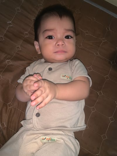 A close-up, eye-level shot captures a baby lying on a brown, diamond-patterned mat, possibly a crib or play mat, in Sungai Meriam, Indonesia. The baby, appearing to be around 6-12 months old, has dark hair and is dressed in a light beige, short-sleeved onesie with two buttons at the neckline and a small, orange and green tag on the left chest. The onesie has a label on the left side of the chest with text that appears to read "Aldo bra M by usita". The baby's hands are clasped together in front of their chest. The background is the brown mat, with a faint shadow on the right side of the frame indicating a light source. The focus is sharp on the baby's face and hands, with a slight blur on the edges.