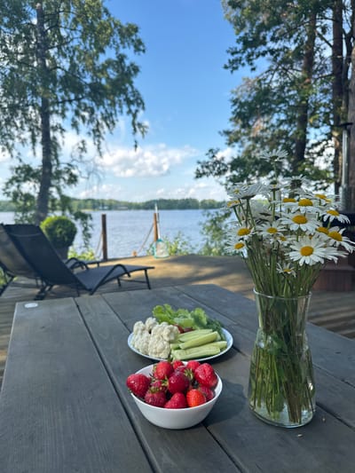 The image depicts a serene outdoor scene at a lakeside location, likely in Priedesmuiža, Latvia. A weathered wooden table is set with a white bowl of fresh strawberries in the foreground and a plate of mixed vegetables, including cauliflower florets, cucumber slices, and lettuce, positioned behind it. To the right of the vegetables, a clear glass vase holds a large bouquet of white daisies with yellow centers.

In the background, a calm lake stretches towards a tree-lined horizon under a bright blue sky with scattered white clouds. Two black lounge chairs are visible on a wooden deck overlooking the water, suggesting a relaxing atmosphere. The overall setting is tranquil and natural, with lush green trees framing the scene. No people are present.