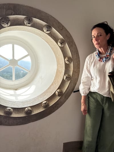 A woman stands next to a circular window in the Quinta da Regaleira, located near Sintra, Portugal. The woman has dark hair pulled back, and she wears a white blouse, green pants, and a patterned scarf around her neck. The circular window has a white frame with six panes, and it is surrounded by a textured stone border decorated with bell-shaped protrusions. The window offers a view of a landscape with trees and hills. The room is lit by natural light, illuminating the beige-painted walls.