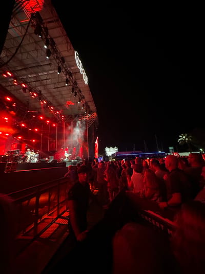 An outdoor concert is taking place at night, with a stage set up under a large canopy structure. Red lights illuminate the stage, casting a dramatic glow on the performers and creating a smoky atmosphere. A band is visible on stage, with musicians playing instruments and a drummer behind a kit. A crowd of people is gathered in front of the stage, watching the performance. The concert appears to be held in a venue near a body of water, with palm trees visible in the background. The lettering "GRAN CASINO" is visible on the canopy. This location is the Gran Casino Costa del Sol in Chiclana de la Frontera, Spain.