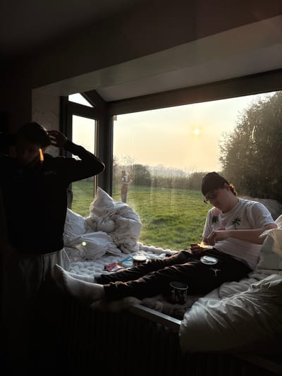 The image depicts an indoor scene within a modern room featuring a large, corner-configured window that overlooks a rural landscape, presumably located in or near the commune of Bû, France. The time of day is characterized by a low sun visible in the pale yellow-orange sky, suggesting late afternoon or early morning.
Three individuals are present. Indoors, on the left, a male in a dark sweatshirt displaying "BROOKLYN" and light-colored trousers stands partially silhouetted against the window, with his hands raised towards his head. To his right, a second male is seated on a window bench piled with white bedding. This individual wears a dark beanie, glasses, a white t-shirt with palm tree motifs, and dark sweatpants with a white Jordan logo. He is engaged in an activity involving a transparent card and a small circular container held in his lap, with a white cup nearby.
Outdoors, visible through the window in the middle distance, a third individual, male, stands on a green grassy field. He is wearing a dark top with a white number "10" on the back and dark trousers. The field is bordered by trees and shrubs under a clear sky. A metal radiator is visible below the window seat indoors.