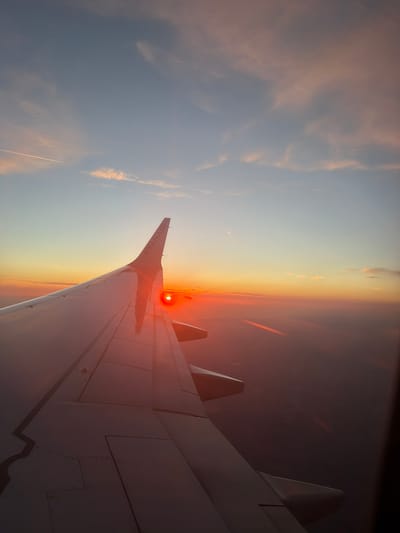 An airplane wing, identified by the "RYANAIR" logo on its tip, is prominently featured in the foreground of an aerial view. The wing, cast in the warm hues of a sunset, extends diagonally across the frame. The sun, a vibrant red orb, is visible just above the horizon, peeking through a band of clouds that separates the sky from the distant land. The sky transitions from a soft blue at the top to a gradient of orange and yellow towards the horizon, with thin, wispy clouds scattered throughout. The terrain below is largely obscured by a hazy, reddish glow from the setting sun, though faint outlines of the landscape, possibly urban or rural, can be discerned. The image captures a moment during a flight, likely over or near Póvoa de Lanhoso, Portugal, given the context provided. There are no people or specific actions depicted, only the static elements of the aircraft and the natural environment.