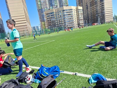 This outdoor photograph captures a sunny day at a soccer field in Novoe Devyatkino, Russia, with residential buildings in the background. The scene depicts what appears to be a youth soccer practice or game.
In the foreground, to the left, a boy wearing a light green "KELME" brand athletic shirt and dark blue shorts stands, holding a dark blue water bottle. He has short brown hair and is looking towards the left. Next to him, on the artificial turf, are several sports bags, including a black one with a yellow draw-string that clearly displays the "SKECHERS." logo. A pair of white and red cleats is visible among the bags. Another boy in a red shirt is partially visible behind these items.
To the right, closer to the midground, another boy with brown hair is seated on the artificial green turf, wearing a blue patterned shirt and dark blue shorts, possibly resting or stretching. Scattered around the foreground are various bags, a water bottle, and other personal items belonging to the players.
In the midground, several other boys, dressed in red, blue, and green jerseys, are actively playing soccer on the artificial turf field, which is marked with white lines. Goalposts are visible further back on the field.
The background is dominated by numerous tall, multi-story residential buildings with various window patterns, suggesting a dense urban or suburban environment. The sky is clear and blue, indicating bright, sunny weather. The overall impression is one of an active, energetic day of sports.